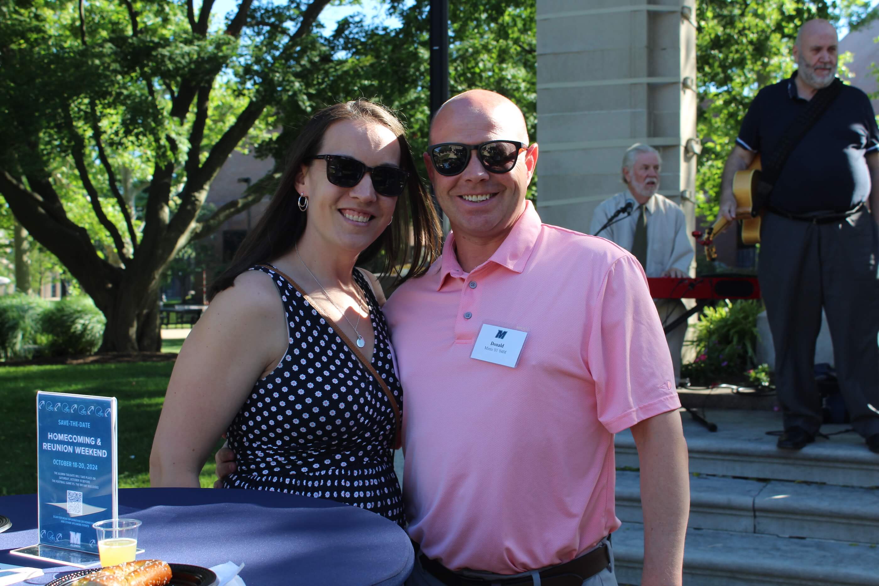 Man and woman, both wearing sunglasses, smiling for camera in front of band