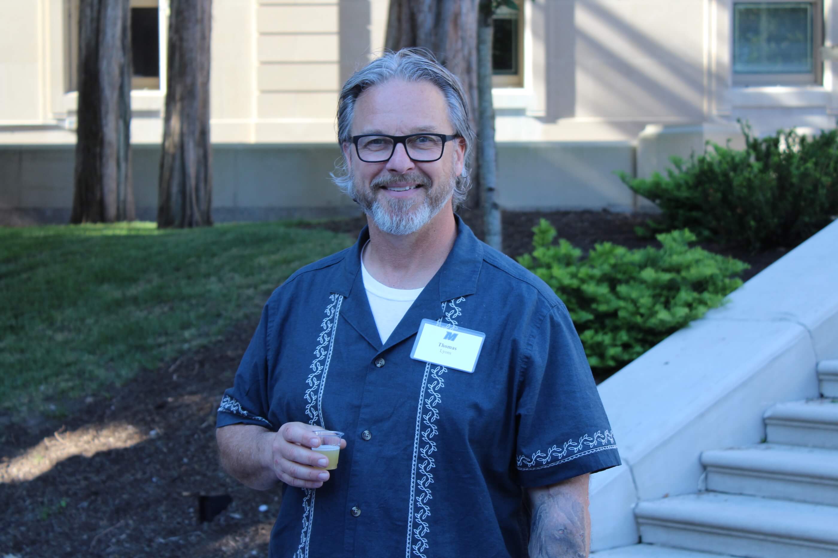 Man in button-up shirt and eyeglasses holding a tasting glass, smiling for camera