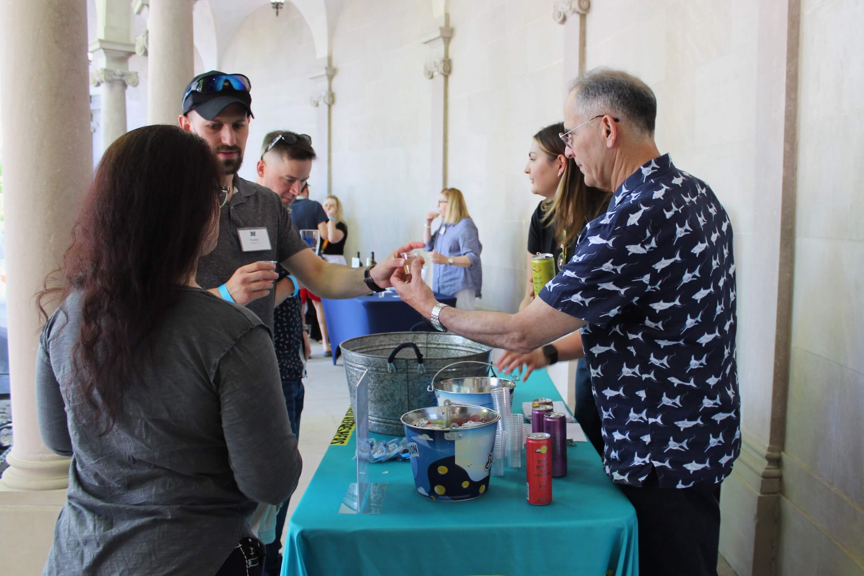 Volunteer in shirt featuring a shark design hands a tasting glass to a man in a baseball cap and sunglasses