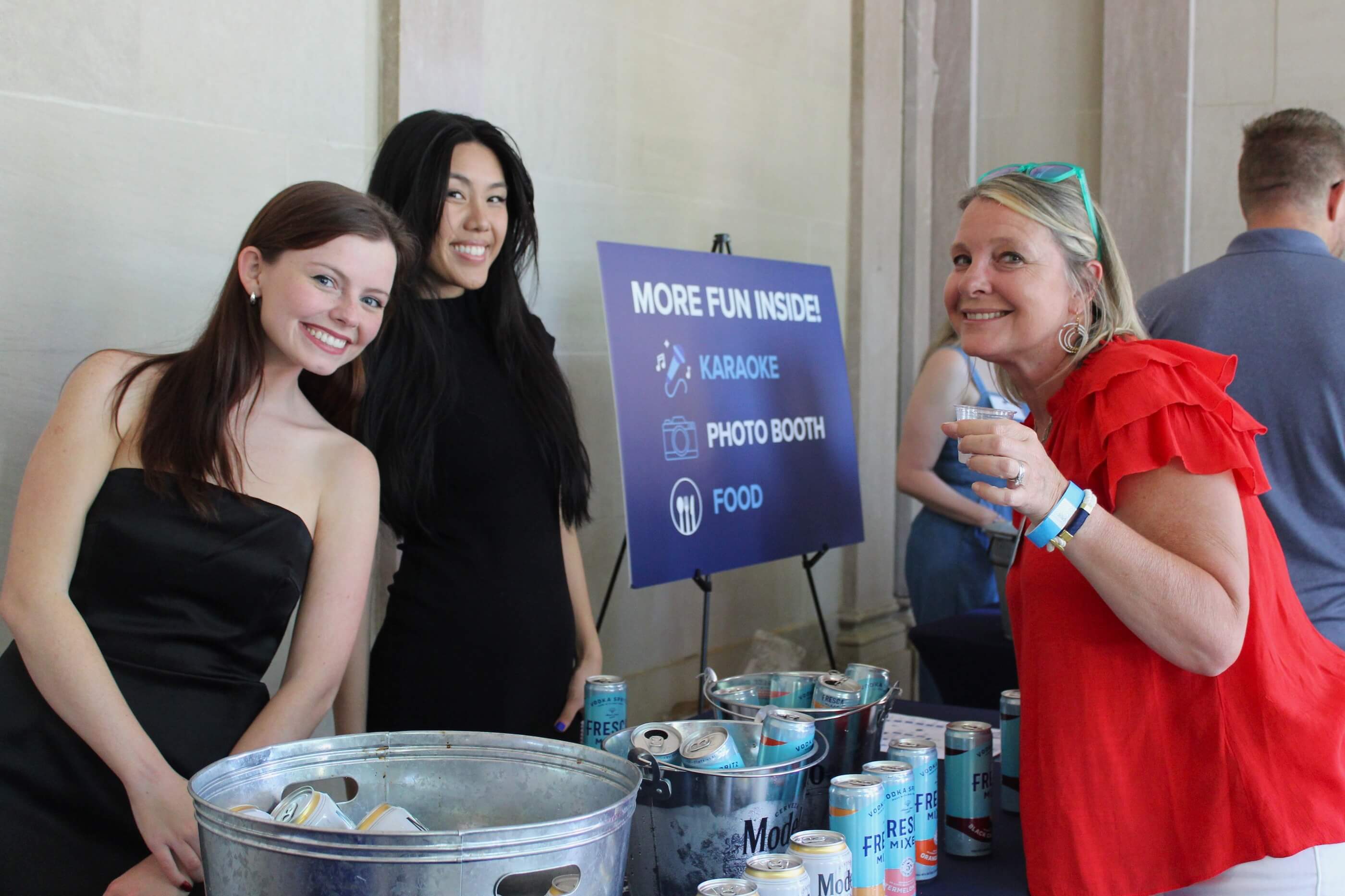 Women posing for a shot near a table that has a sign reading "More Fun Inside: Karaoke, Photo Booth, Food"