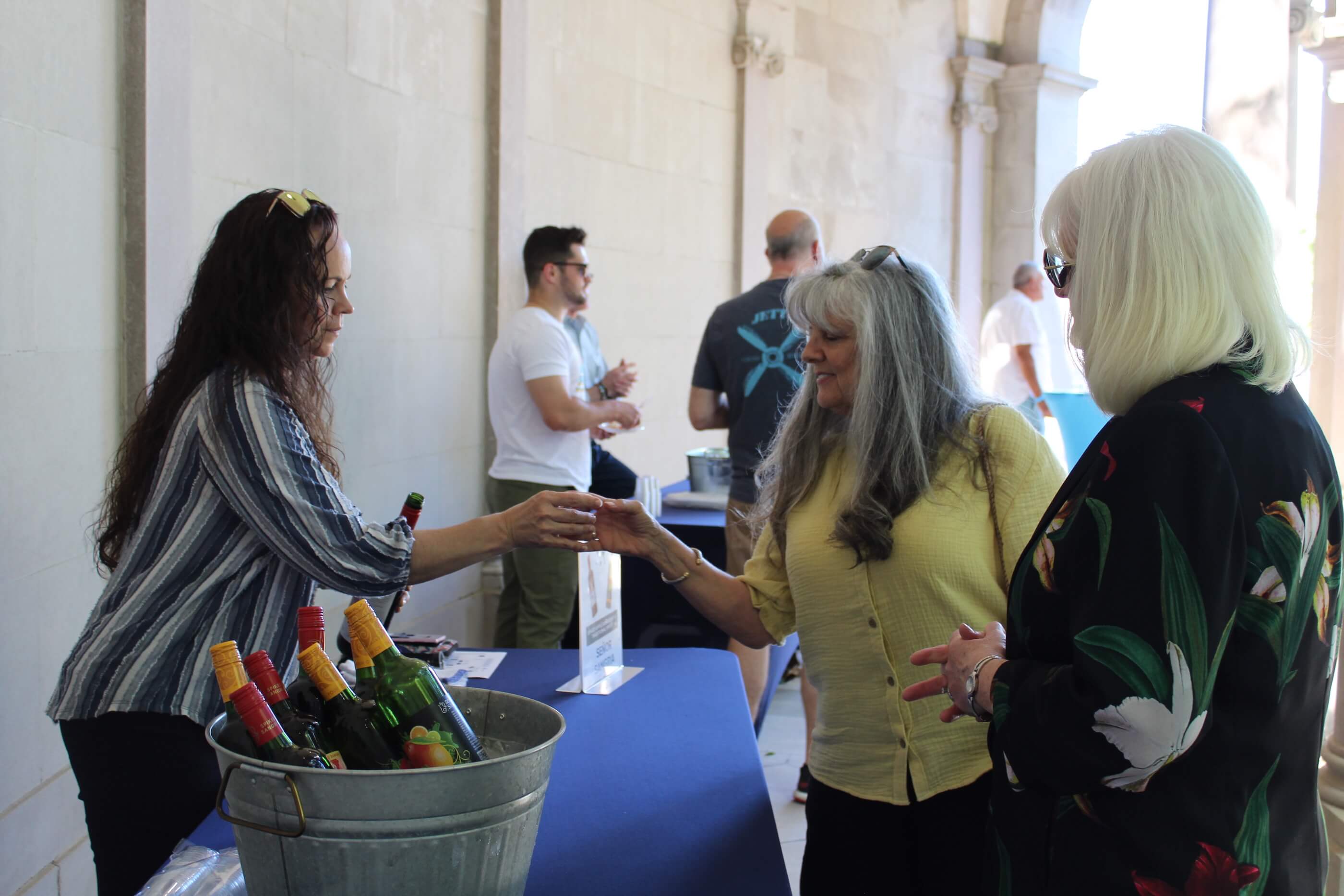 A volunteer hands a woman a tasting glass with red wine as her friend looks on.