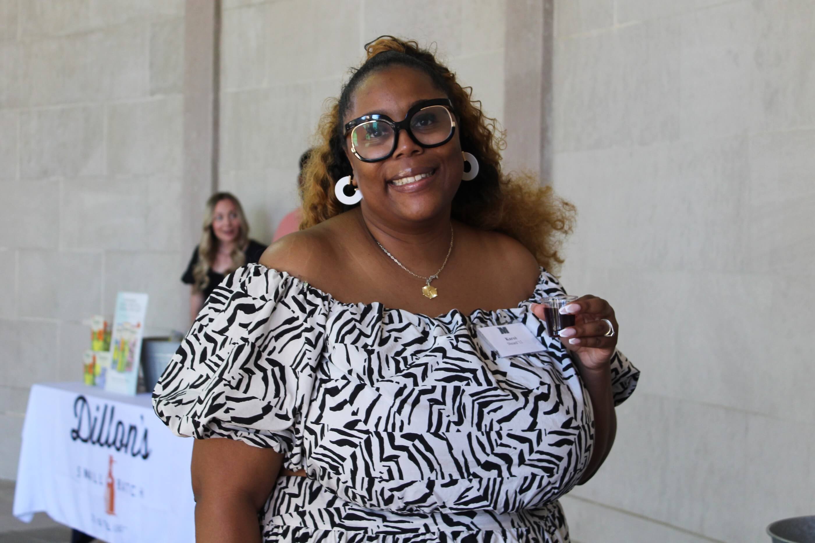 Woman in dress and wearing large earings, posing for a photo in front of the Dillon's Small Batch Distillers table