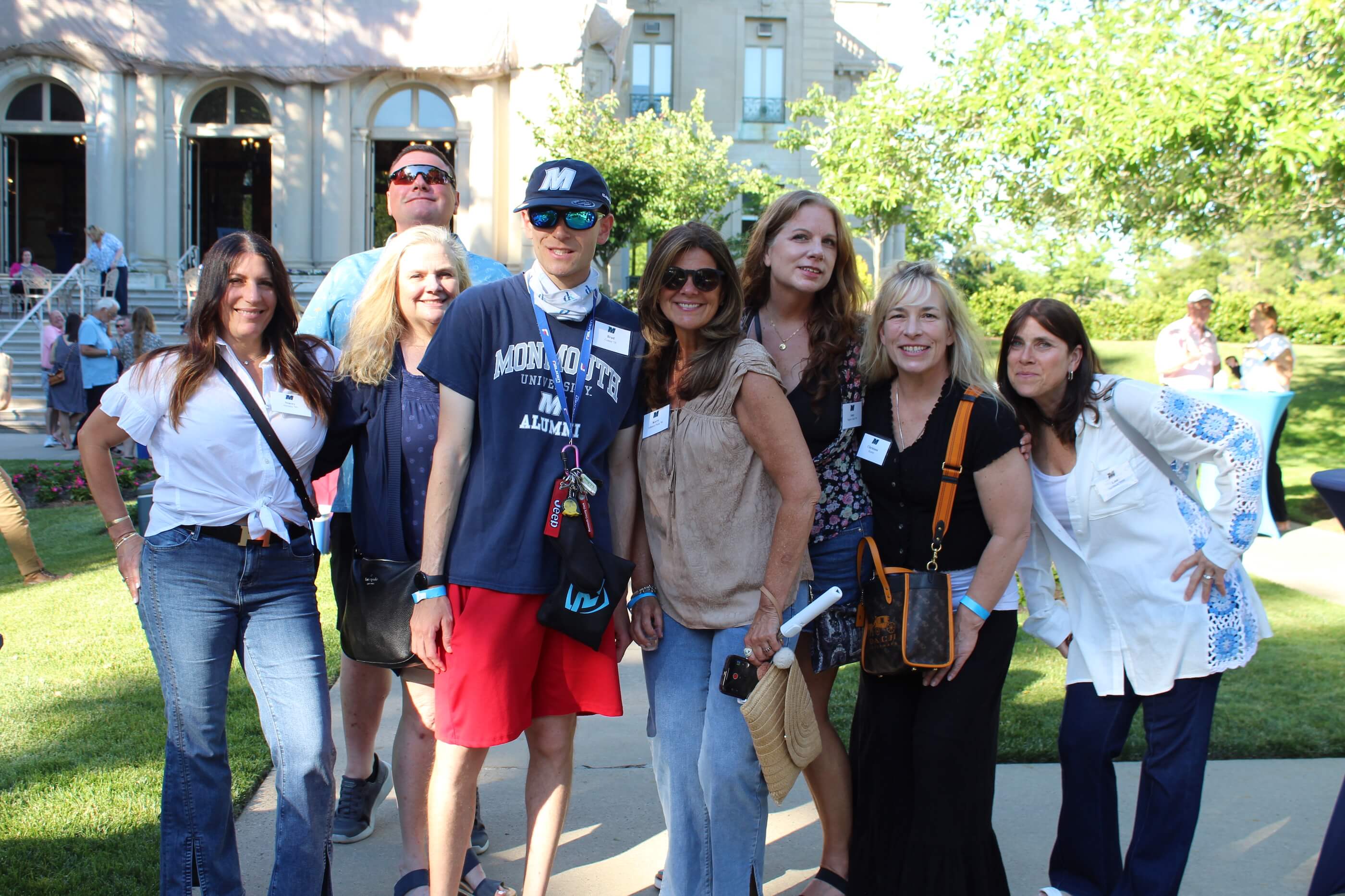 Eight people posing together for a group shot, some wearing Monmouth regalia. The Great Hall is visible in the background