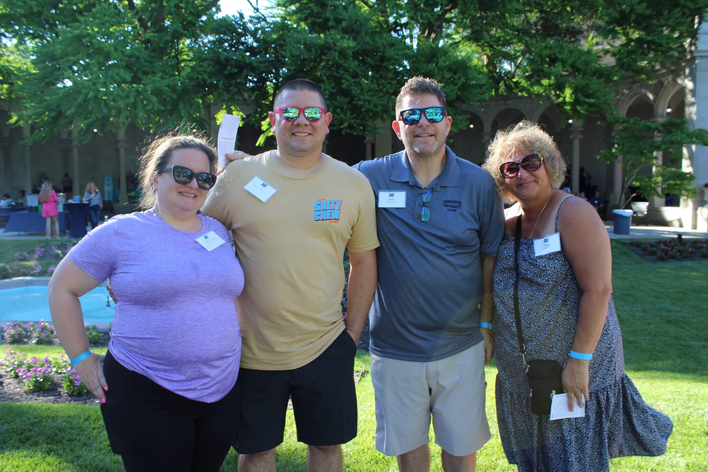 Four attendees wearing sunglasses smiling for the camera, with the green trees of Erlanger Gardens in the background