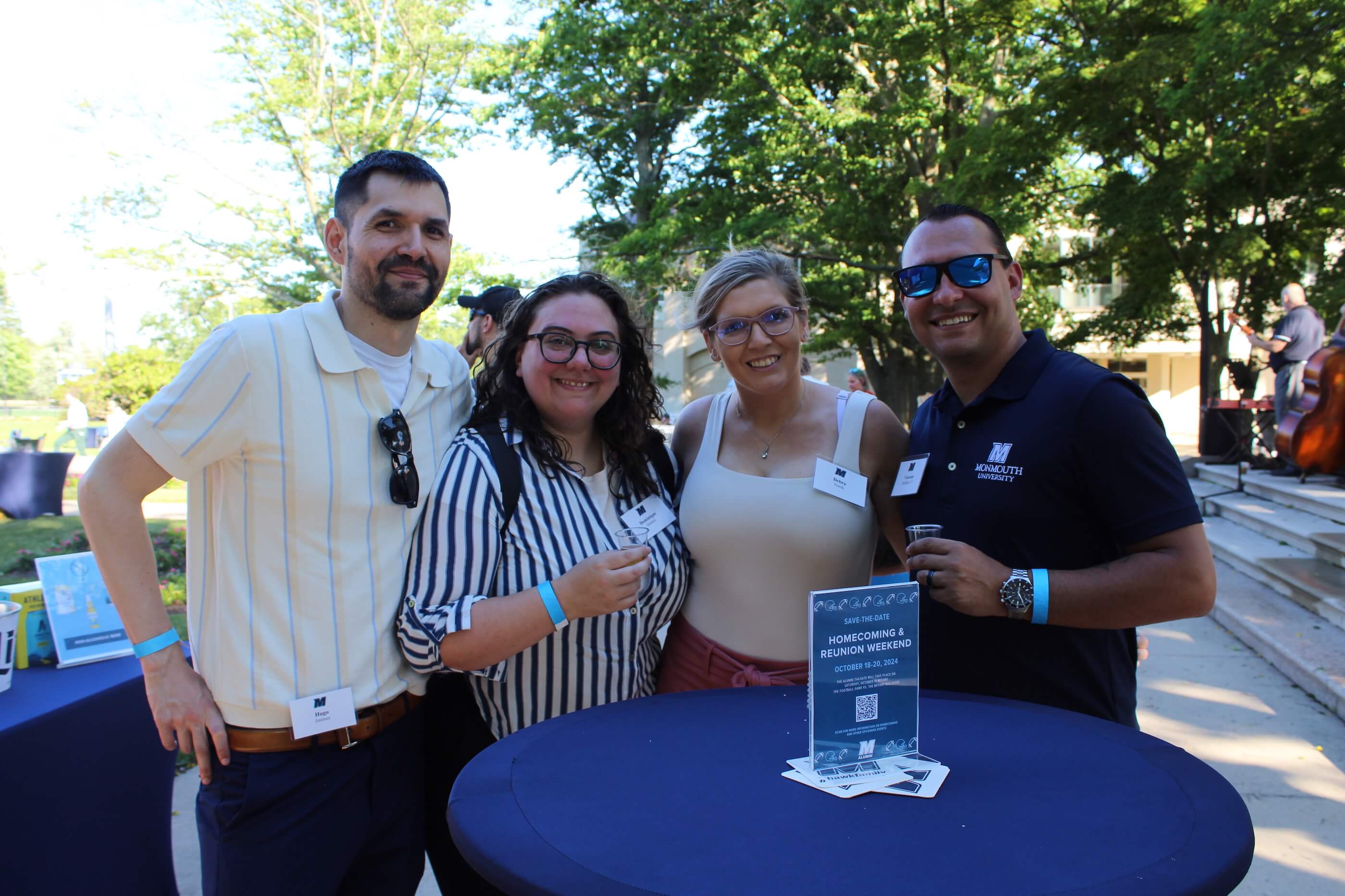 Four attendees smiling for a group shot, two holding tasting glasses, while standing in front of a table. the table has a sign advertising Homecoming and Reunion Weekend