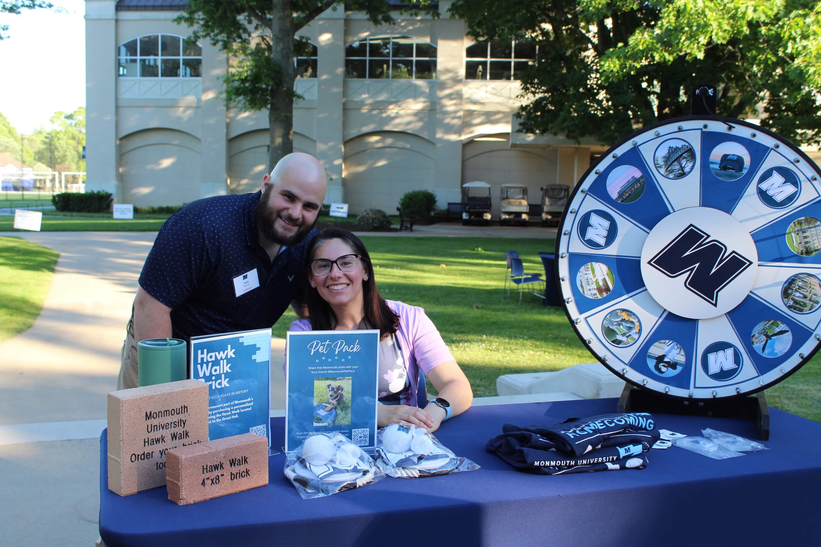 Two tablers posing for a photo by the Hawk Walk Brick campaign table. A spinner picker with the Monmouth logo in the middle sists on the table