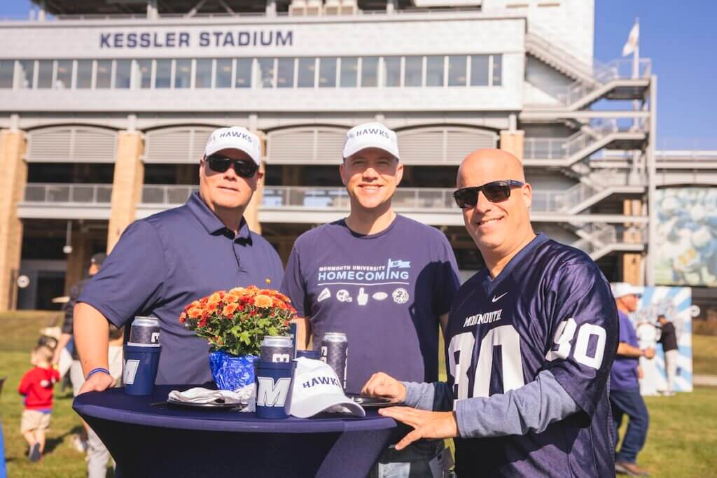 Three men smile for camera in front of stadium by high top table