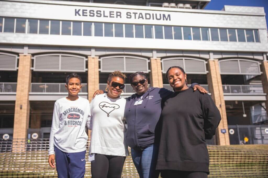 Group of women holding each others shoulders, smiling for camera outside stadium