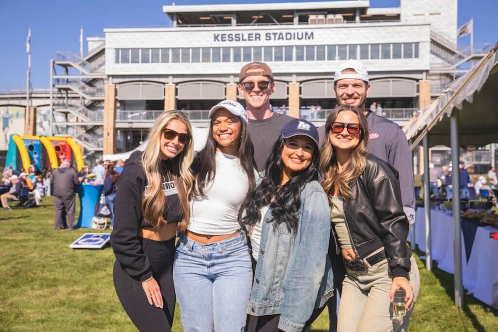 Six alumni in monmouth gear pose for photo in front of stadium