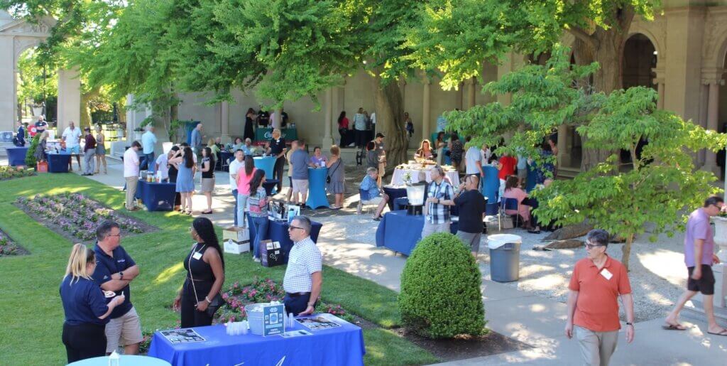 A crowd of wine and stein attendees gather among each other by the fountains, trees and columns of Erlanger Gardens