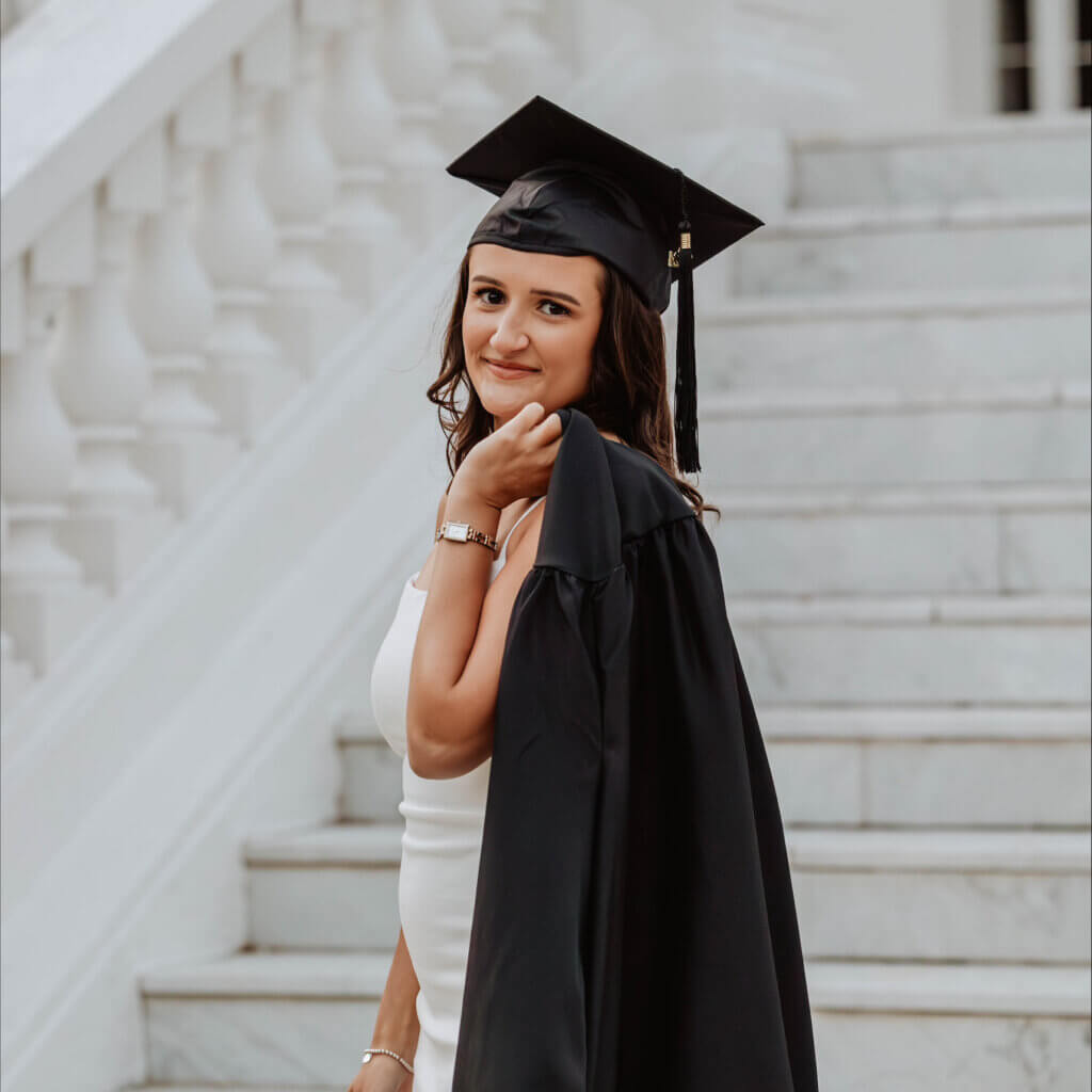 Woman on steps wearing graduation cap, holding her graduation gown over her shoulder