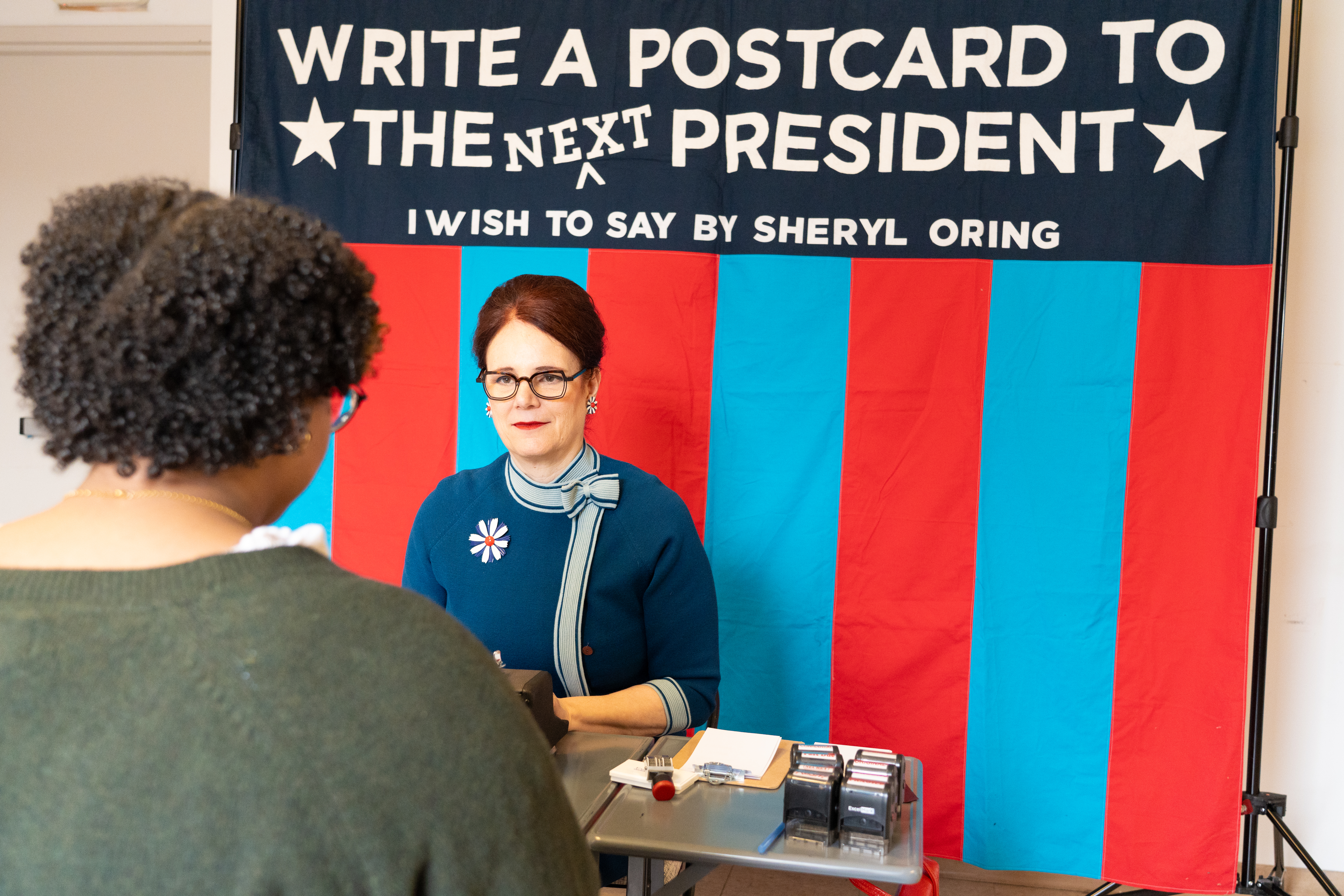 Woman at a table accepting postcards. Behind her, a sign that reads "write a postcard to the next president"