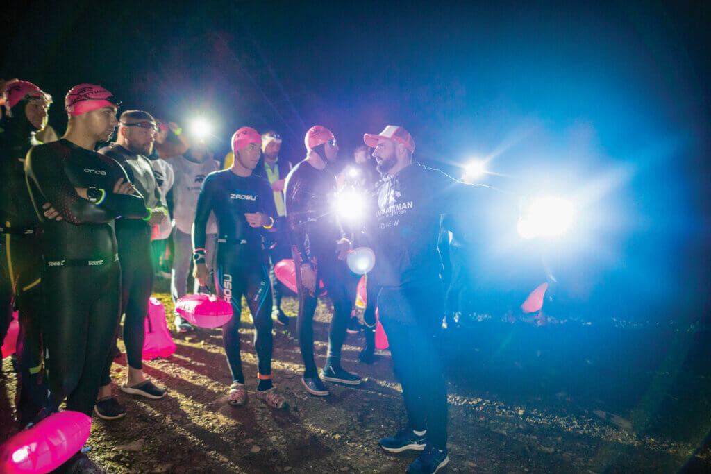 Triathletes in swimsuit, some with headlamps, gather before the race.
