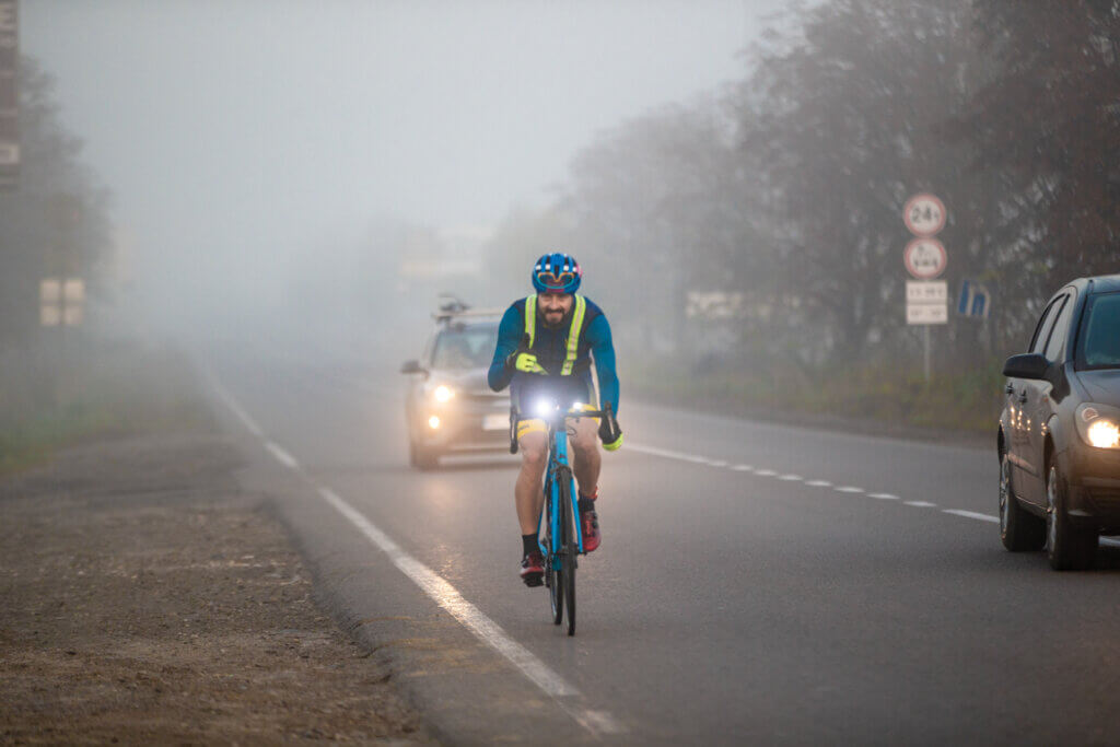 A triathlete biking along a foggy road, trailed by his trail car.