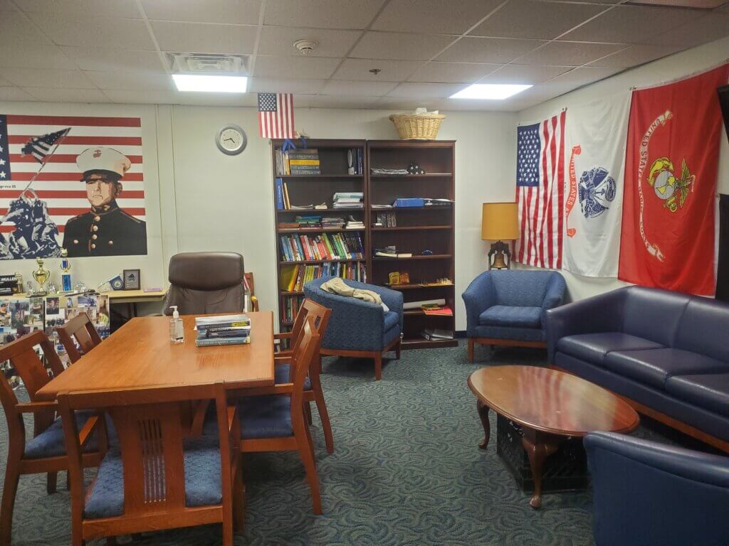 Inside Student Veterans Lounge is a table, with couches, and a bookshelf pictured. Mural of LCpl Cosgrove also pictured.