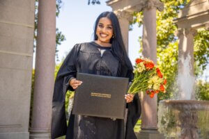 A smiling graduate in cap and gown holds a diploma and bouquet of flowers at Erlanger Gardens