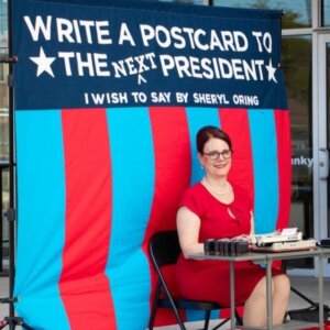 Sheryl Oring seated at typewriter in front of flag with words, Write a postcard to the next president"