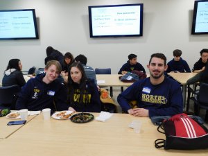 3 Toms River North students, 1 female in between 2 males, smiling and eating lunch in front of a group of high school students