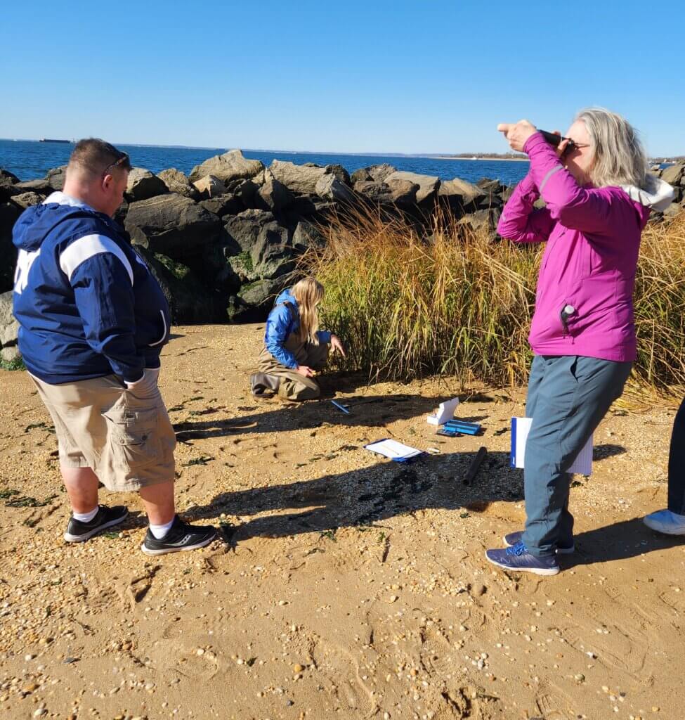 Woman taking photos on a beach while two other people survey the area. Beach rocks and weeds in the background