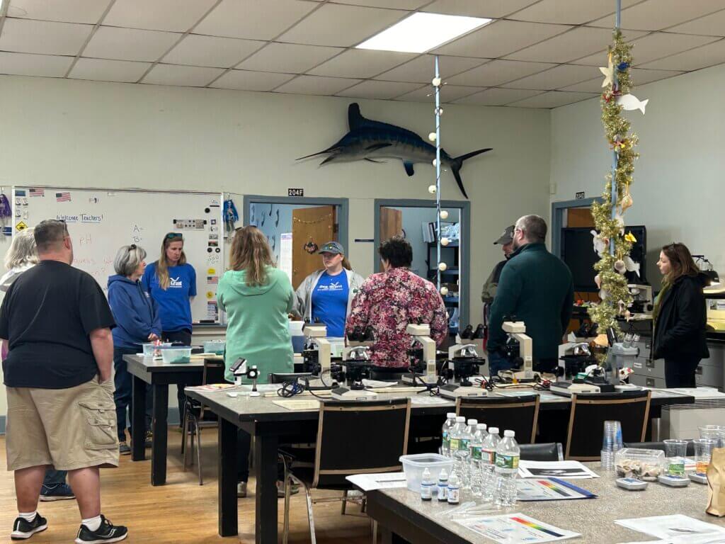 A woman speaks to an attent group of adults in a classroom. Scientific microscopes and plastic containers sit on the tables. A swordfish hangs off one of the walls. 