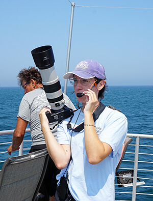 Brooke van de Sande stands aboard a vessel, speaking through a headset mic while holding a camera.